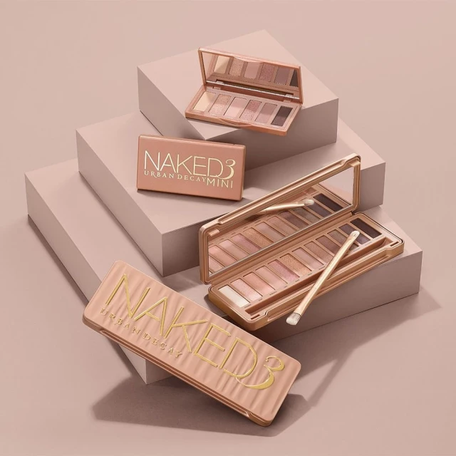 Still life studio shot featuring multiple Naked3 Urban Decay eyeshadow palettes in a warm, neutral color scheme on stepped platforms.