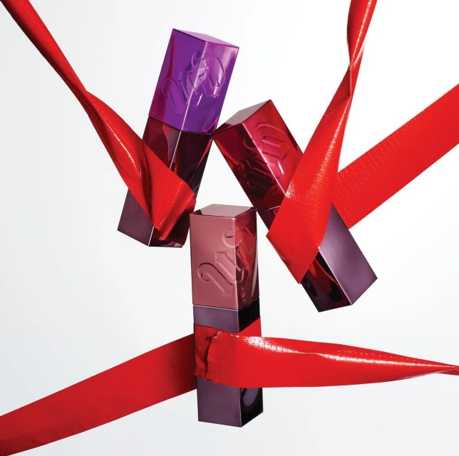 Three Urban Decay lipsticks with purple and pink packaging are adorned with shiny red ribbon against a bright white background.