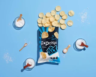 Top-down view of a Popchips Sea Salt bag surrounded by loose crisps, small wooden spoons, and salt bowls on a blue surface.
