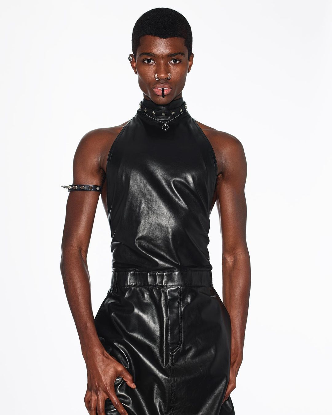 Man in black leather halter top and pants poses in a fashion shot against a white backdrop wearing spiked collar and arm cuff.