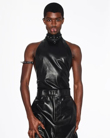 Man in black leather halter top and pants poses in a fashion shot against a white backdrop wearing spiked collar and arm cuff.