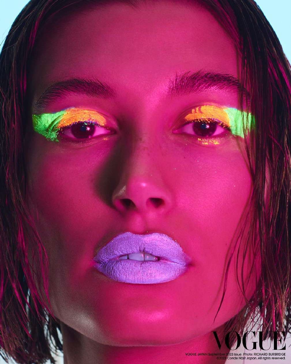 Portrait of a woman with colorful eyeshadow and lipstick. The colors are neon green, yellow, pink, and purple against a blue background.