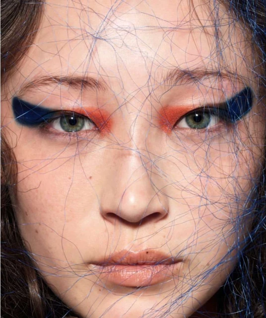 Close-up portrait of a woman with dramatic eye makeup, featuring orange eyeshadow and bold blue eyeliner, with fine blue strands overlaid.