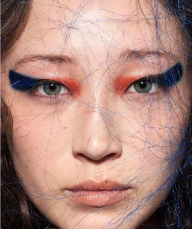 Close-up portrait of a woman with dramatic eye makeup, featuring orange eyeshadow and bold blue eyeliner, with fine blue strands overlaid.