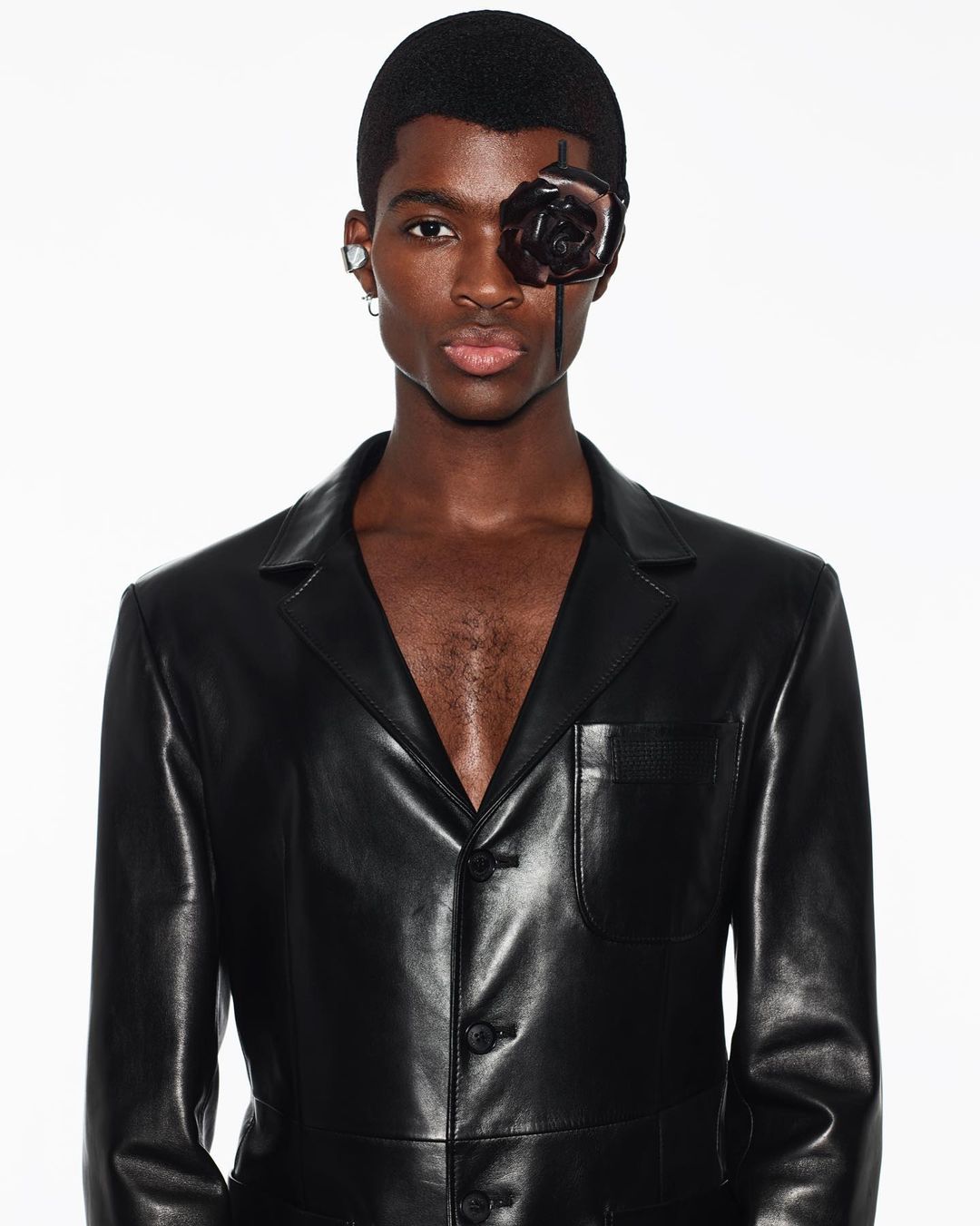 Portrait of a dark-skinned man wearing a black leather jacket and a black rose eye accessory against a white background in a photo studio.
