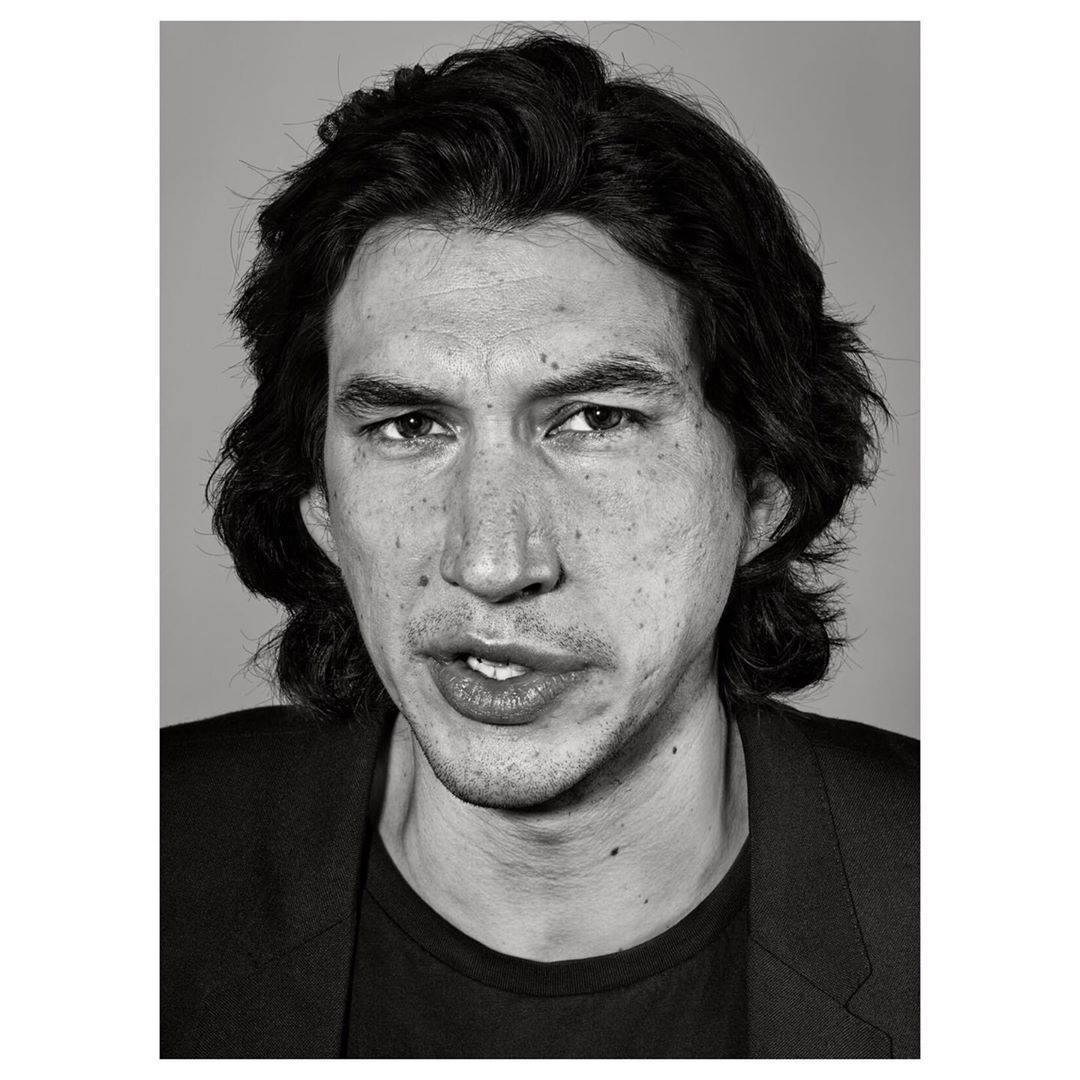 Black and white headshot of actor Adam Driver in a dark jacket against a grey backdrop, capturing a serious yet slightly wry expression.