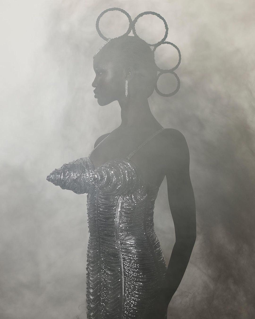 Portrait of a Black woman in a shimmering silver dress and headpiece, looking left against a smoky background, long earrings.