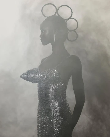 Portrait of a Black woman in a shimmering silver dress and headpiece, looking left against a smoky background, long earrings.