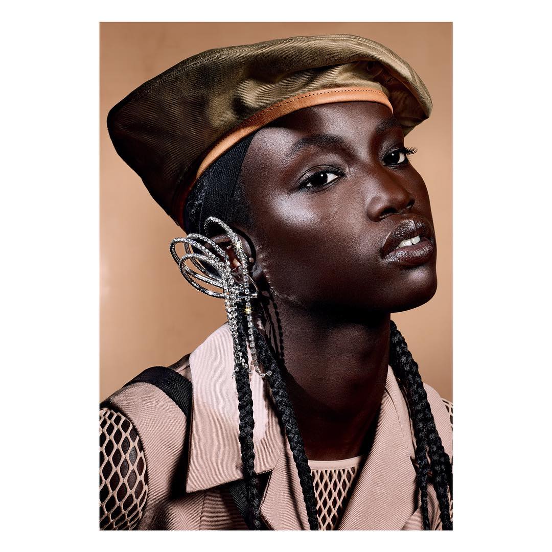 Close-up portrait of a Black woman in a tan beret and large sparkling earring, wearing a tan and black top, against a tan background.