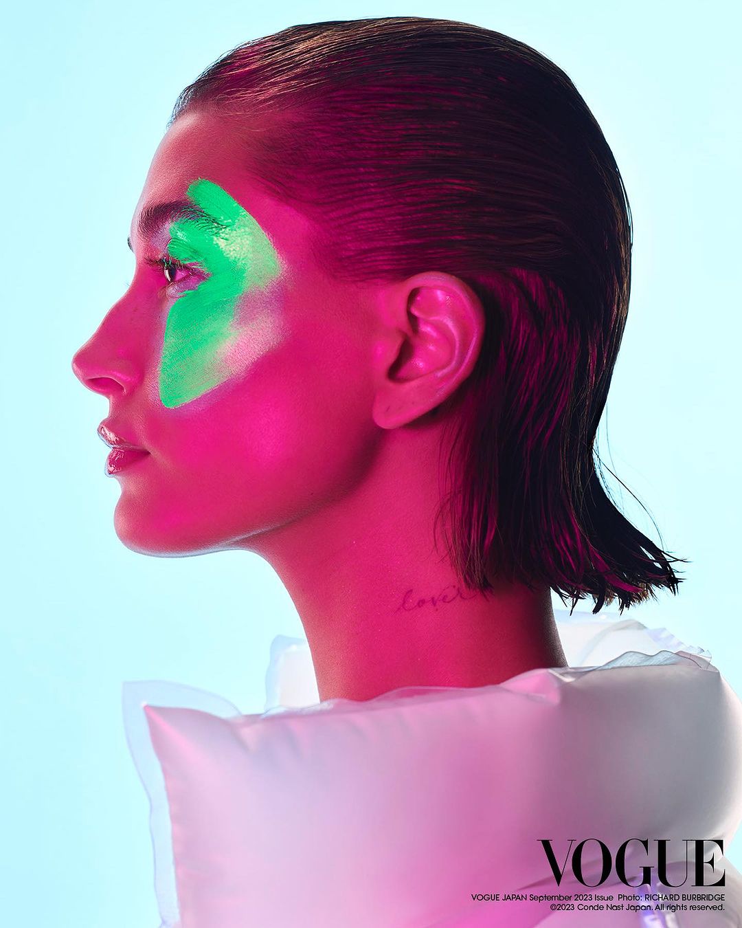 Close-up studio portrait of a woman with vivid pink lighting and green face paint in a side profile view against a blue background.