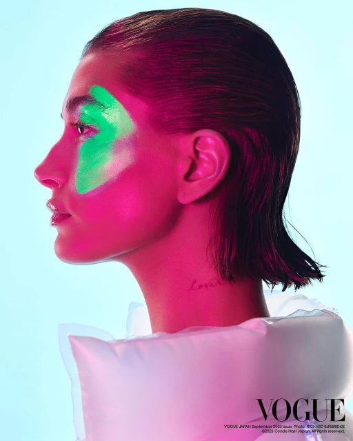 Close-up studio portrait of a woman with vivid pink lighting and green face paint in a side profile view against a blue background.