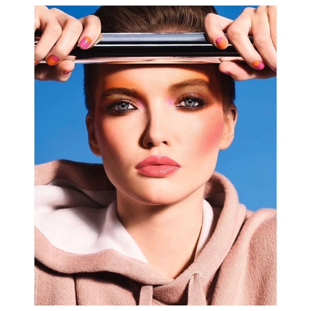 Close-up of a young woman with creative orange and pink eye and nail makeup, wearing a hoodie against a blue background.