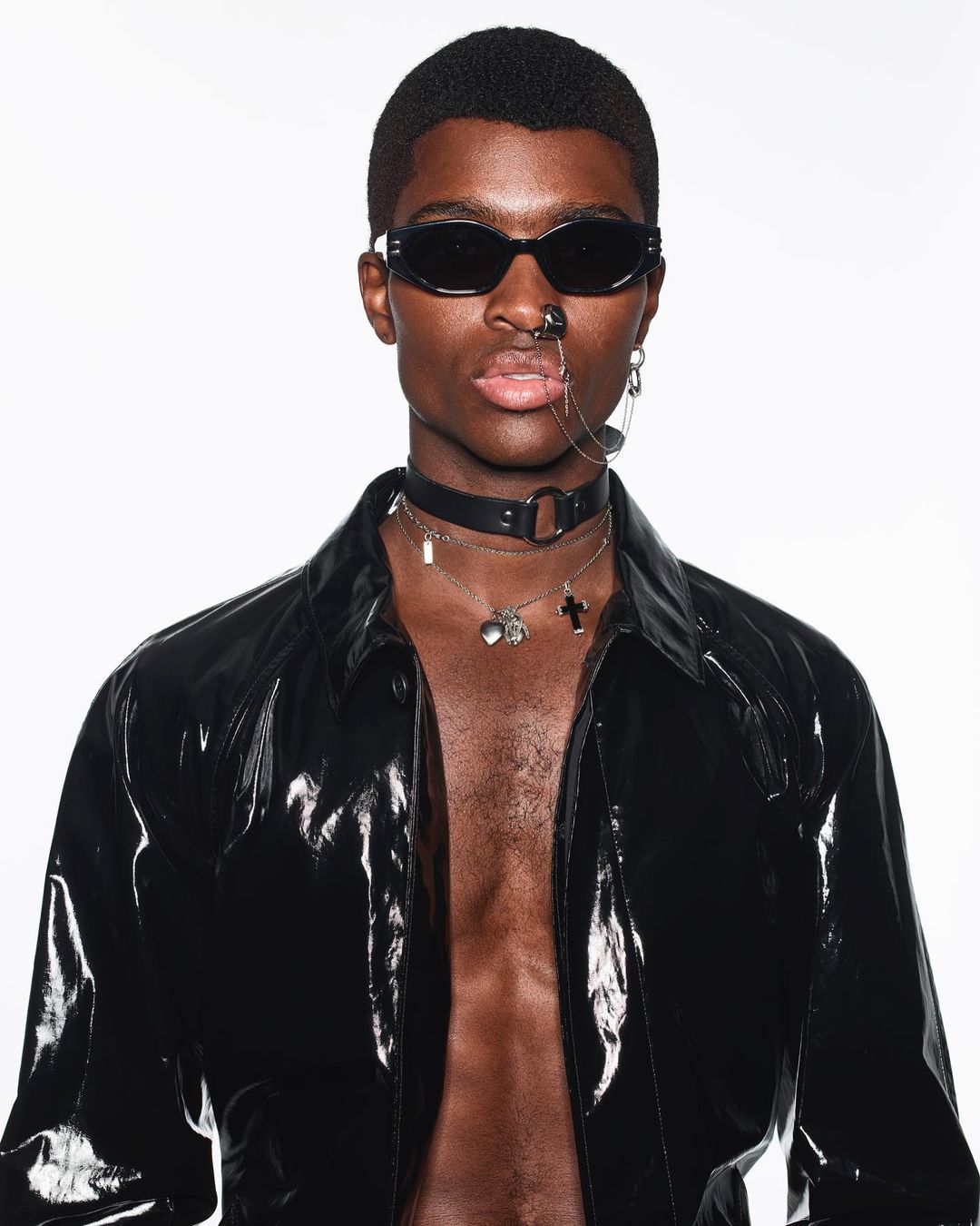 Dark-skinned adult male in sunglasses, open black vinyl jacket, and choker against a white backdrop in a fashion forward close-up studio portrait.