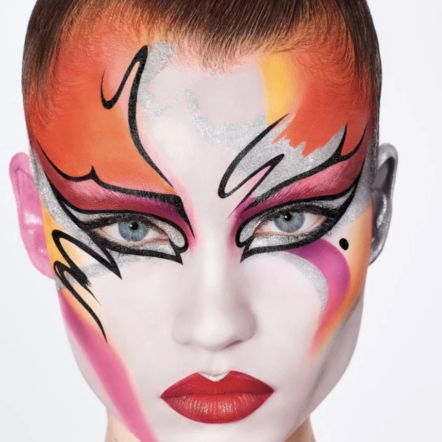 Artistic portrait of a person with red lips and creative makeup using orange, pink, silver, and black set against a white background.