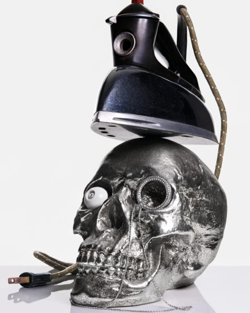 Studio shot featuring a chrome skull topped with a vintage iron; a monocle and white eyeball add a surreal touch against a white backdrop.