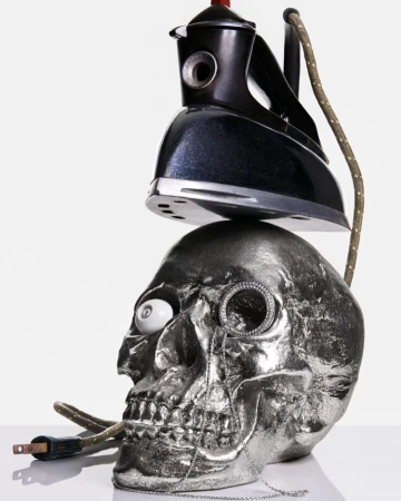 Studio shot featuring a chrome skull topped with a vintage iron; a monocle and white eyeball add a surreal touch against a white backdrop.