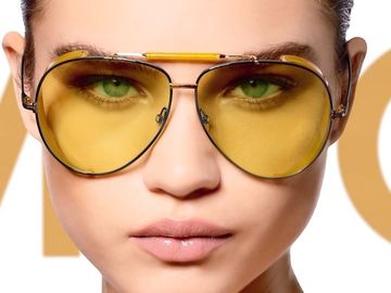 Close-up studio shot of a woman with green eyes wearing yellow tinted aviator sunglasses on a white background.