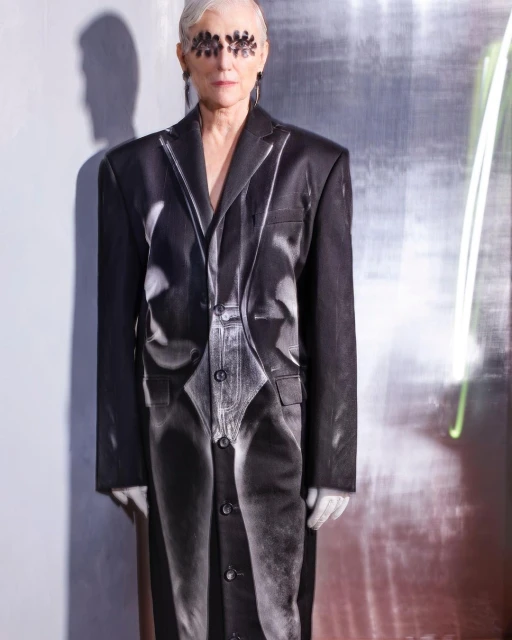 Mature woman with gray hair wearing black eye decor and a printed suit stands indoors with a silver reflective background and neon lighting.
