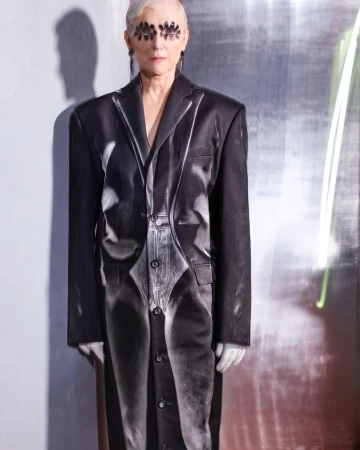 Mature woman with gray hair wearing black eye decor and a printed suit stands indoors with a silver reflective background and neon lighting.