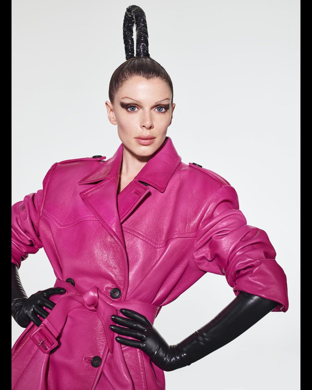 Adult female with dark eye makeup wearing a pink leather trench coat and black gloves against a white backdrop.