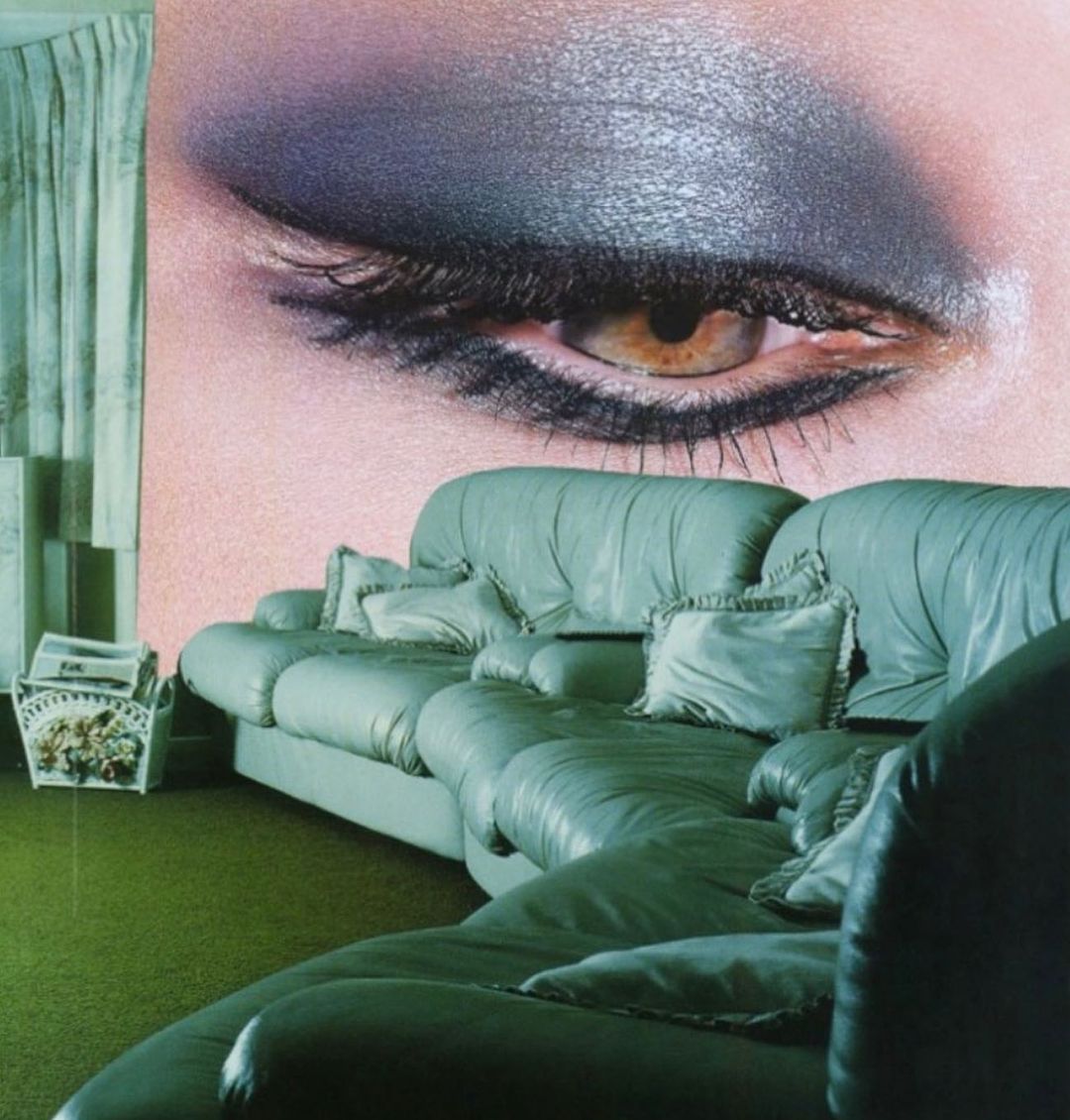 Close-up of a woman's eye with grey eyeshadow on a pastel background, with a green couch, pillows and a basket in the foreground.