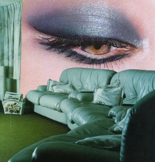 Close-up of a woman's eye with grey eyeshadow on a pastel background, with a green couch, pillows and a basket in the foreground.