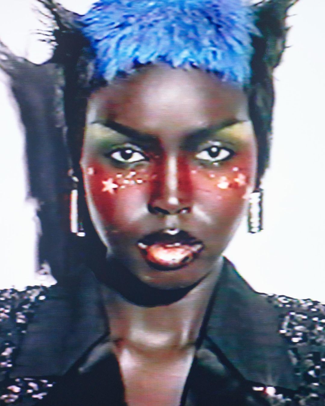 Close-up portrait of a Black woman with blue hair, dramatic green and red makeup, wearing a black sequined top, against a white background.