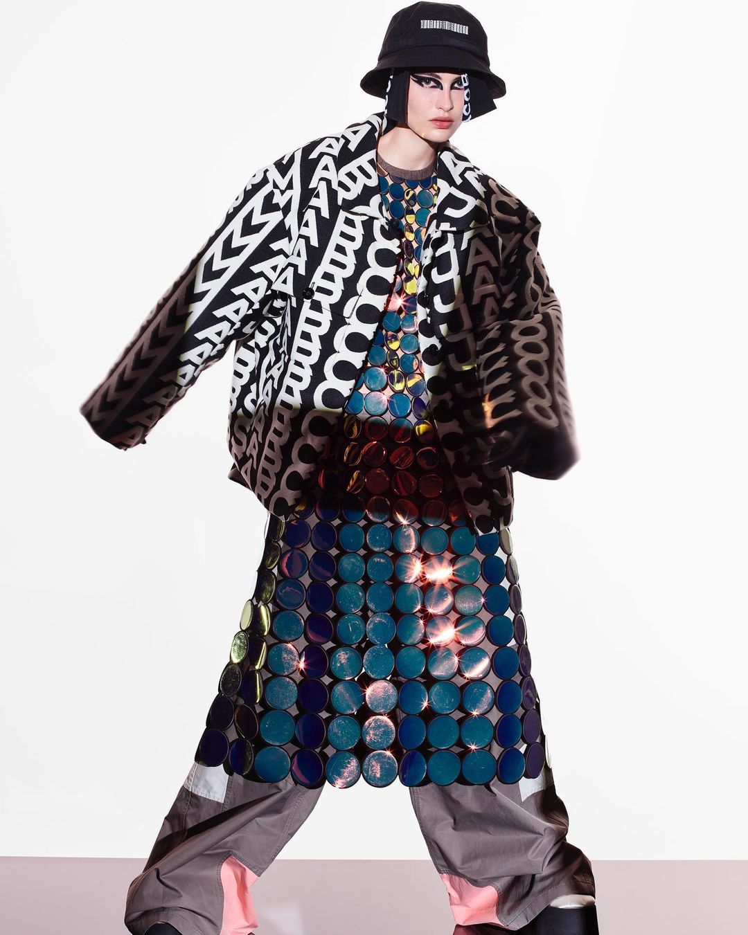 A person in a black and white graphic jacket, bucket hat, a skirt made of blue circles, and gray pants, standing in a white space.