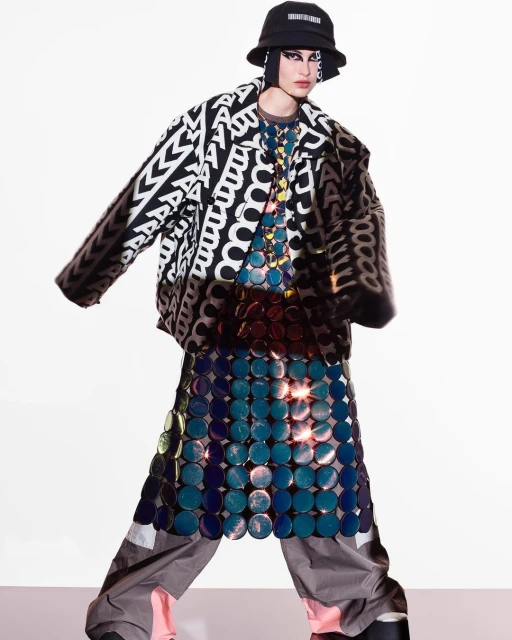 A person in a black and white graphic jacket, bucket hat, a skirt made of blue circles, and gray pants, standing in a white space.