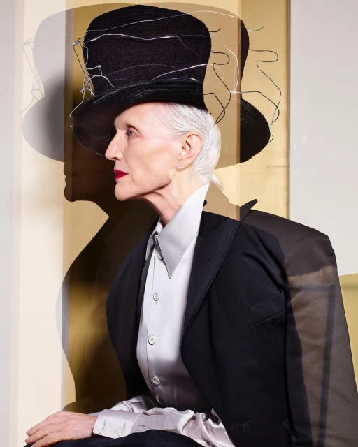 Elderly woman in profile wearing a top hat and black blazer against a neutral background with a shadowy figure.