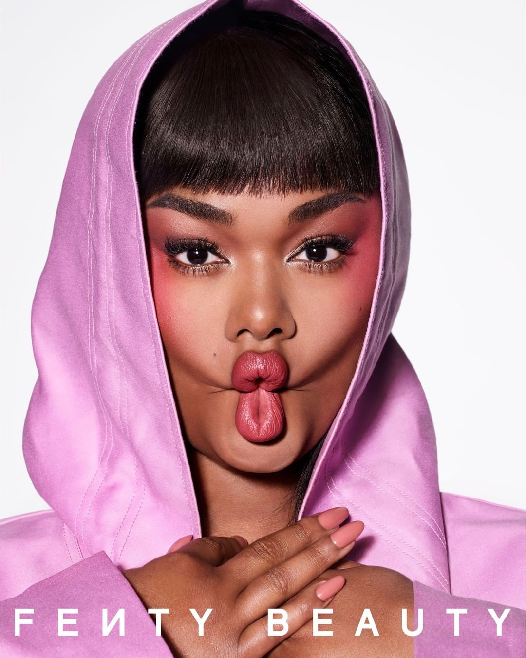 Close-up studio shot of an adult woman with dark hair and pink makeup, wearing a pink hooded top, making a kissy face at the camera.