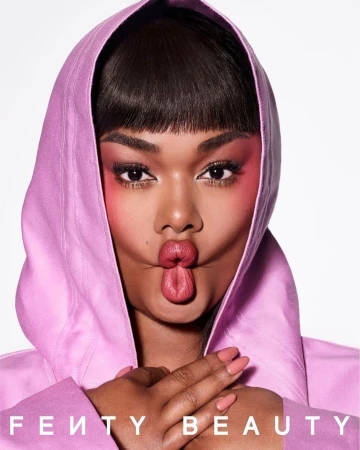 Close-up studio shot of an adult woman with dark hair and pink makeup, wearing a pink hooded top, making a kissy face at the camera.
