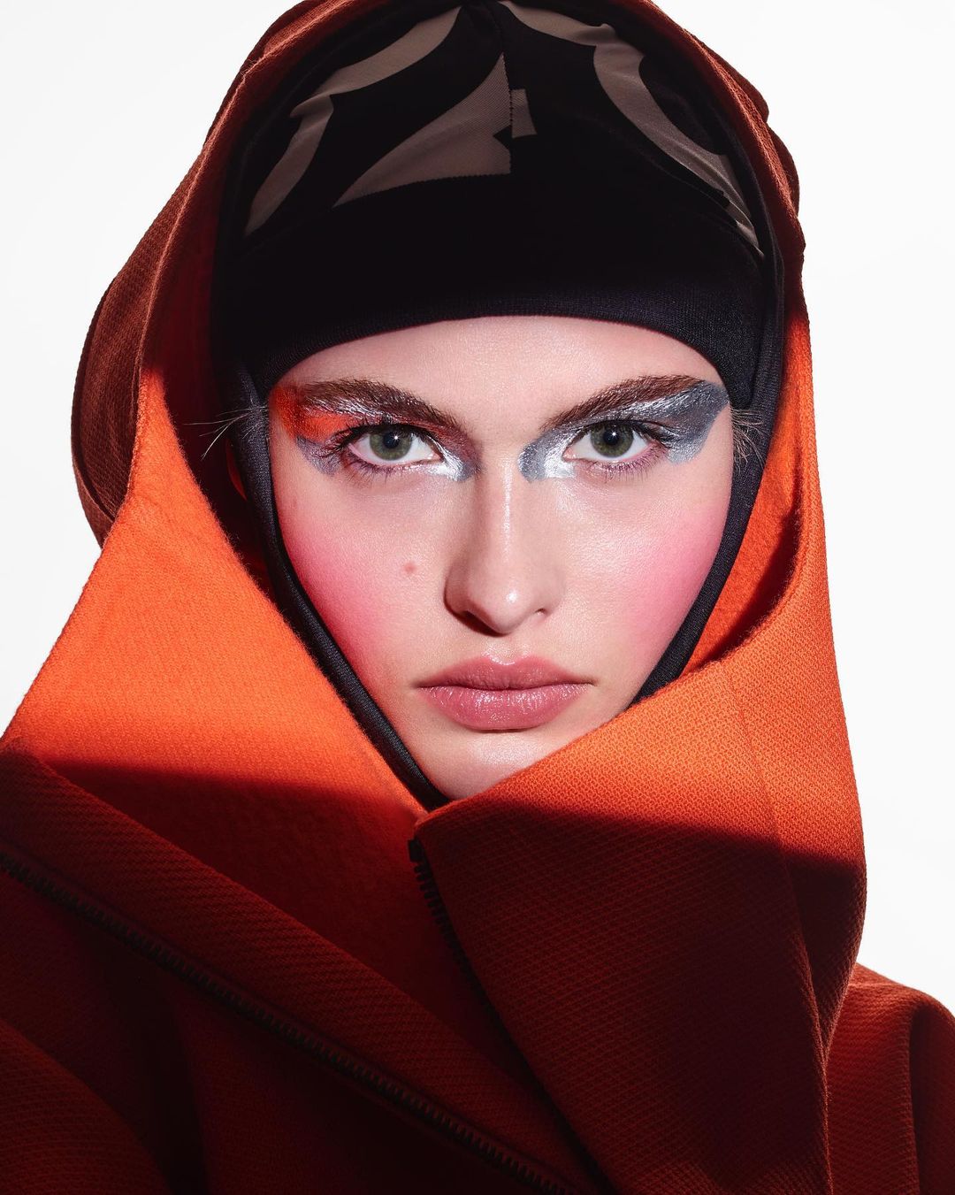 Close-up portrait of a model with bold makeup, an orange hooded garment, and a black patterned head covering against a white backdrop.