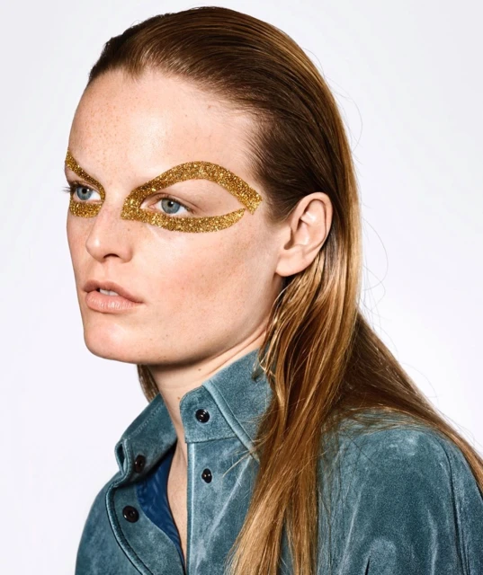 Close-up of an adult female with blonde hair and blue eyes wearing gold glitter eye makeup against a white background.