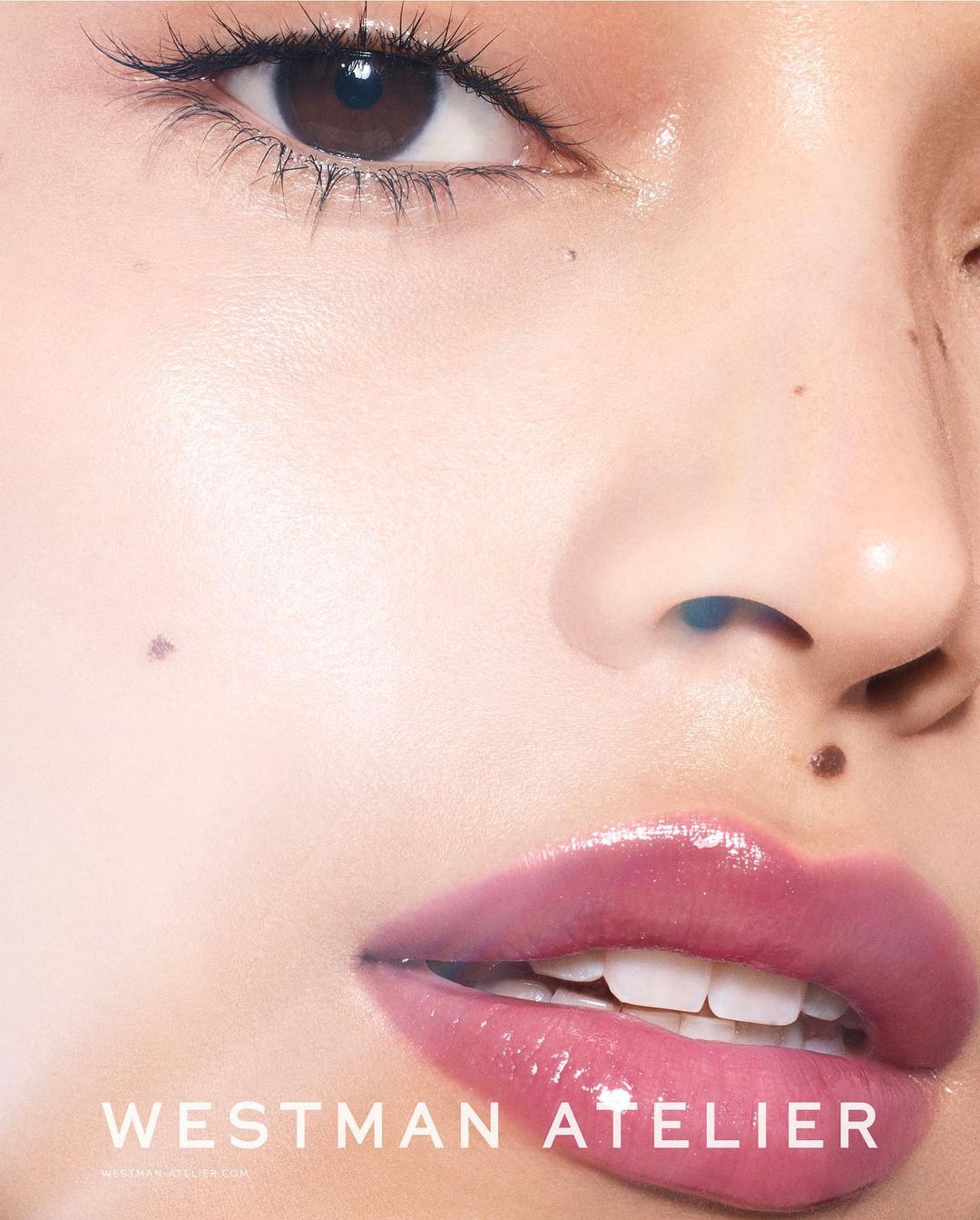 Close-up beauty shot of a woman's face featuring glossy pink lips, fair skin, and dark eyes, showcasing the 'Westman Atelier' brand.
