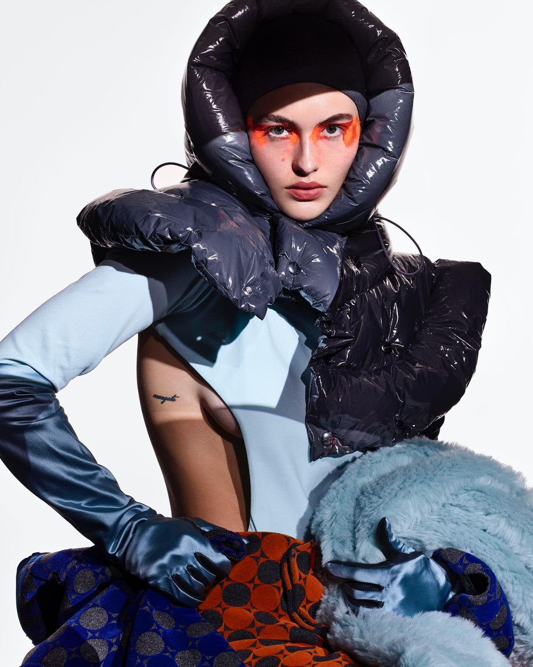 High-fashion portrait of a woman in avant-garde headwear and a light blue bodysuit, makeup around the eyes, gloves, and patterned fabrics.