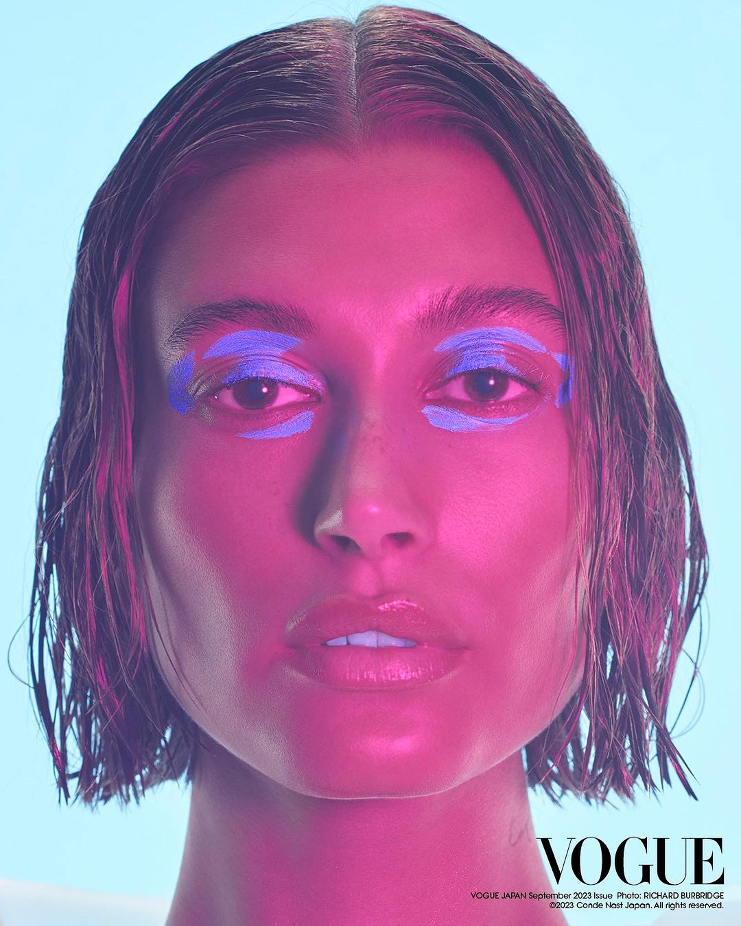 Close-up portrait of a woman with blue eyeshadow and glossy lips against a light blue background, illuminated by pink lighting.