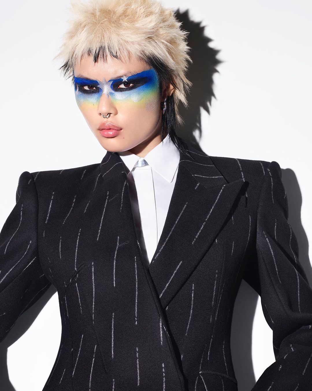 Woman with vibrant makeup and striking blonde hair poses in a pinstripe blazer with a white collared shirt, set against a clean white backdrop.