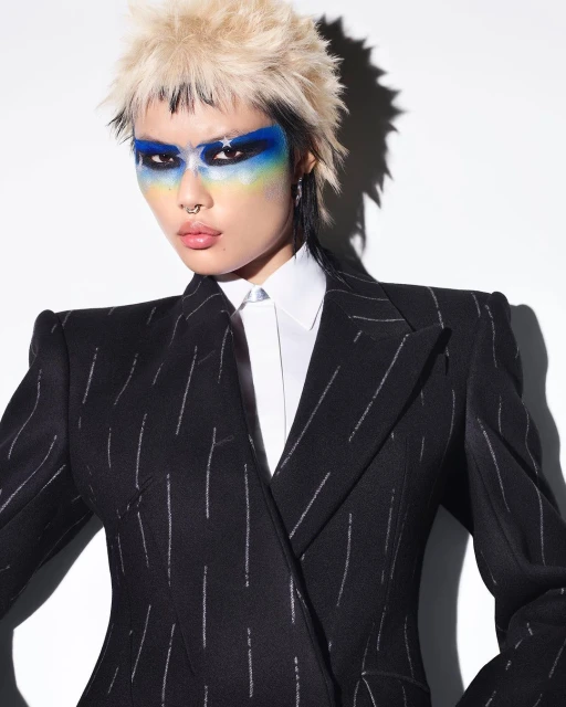 Woman with vibrant makeup and striking blonde hair poses in a pinstripe blazer with a white collared shirt, set against a clean white backdrop.