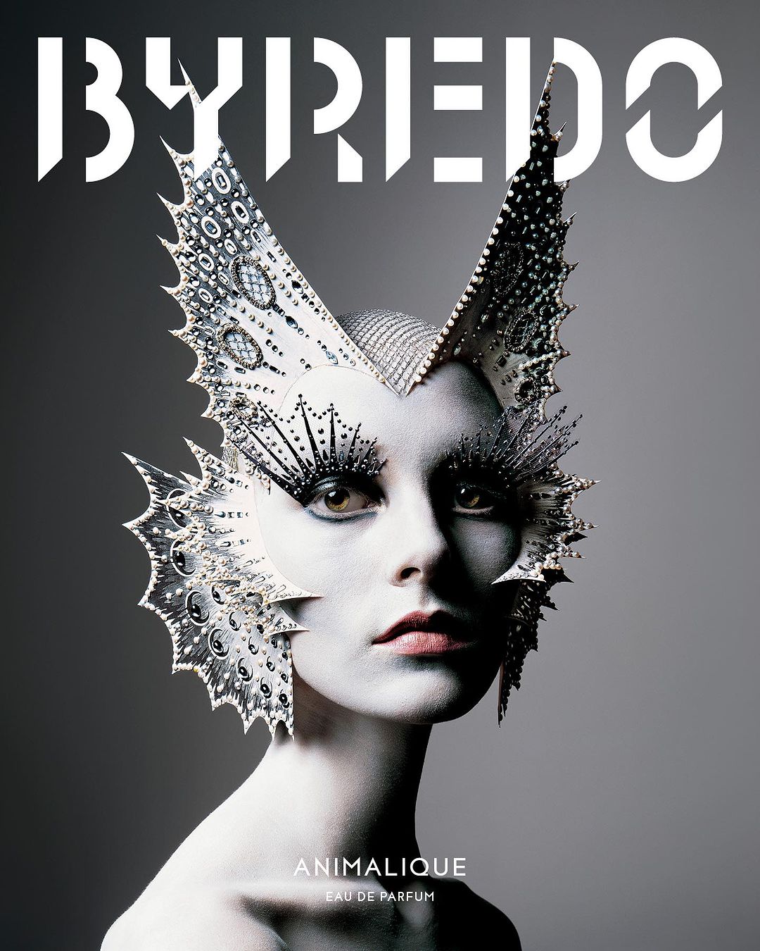 Close-up studio shot of a model wearing an elaborate silver headdress, dramatic eyelash extensions and makeup with a grey background.