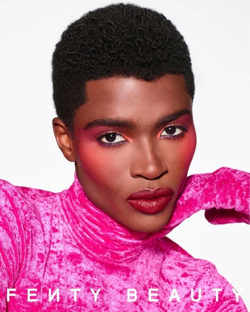 Close-up of a Black male model with pink makeup and a pink velvet top, showcasing Fenty Beauty makeup products against a white background.