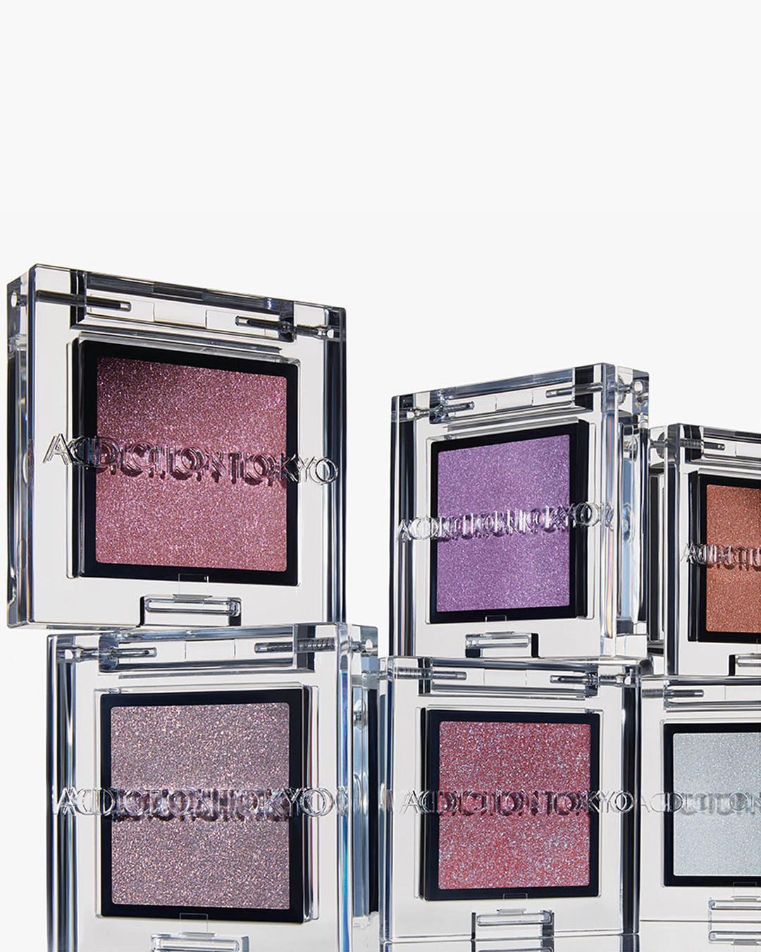 Close-up of stacked ADDICTION TOKYO eye shadow singles in clear cases showcasing pink, purple, and bronze-toned shimmery eyeshadows.