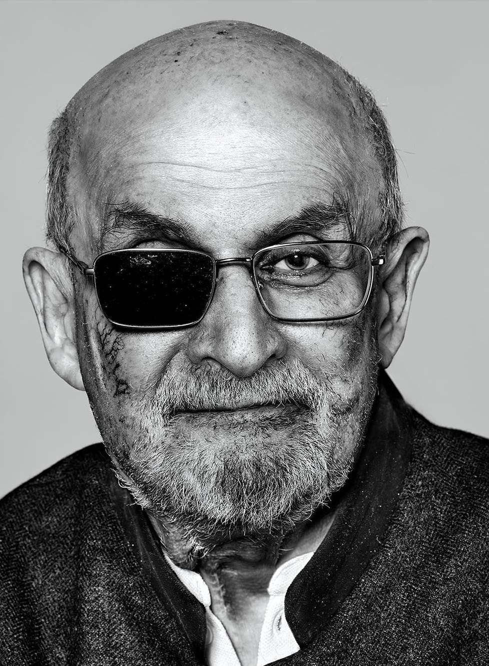 Portrait of an adult male with eyeglasses, including one lens covered by a black patch, with a beard, wearing a dark jacket against a grey background.
