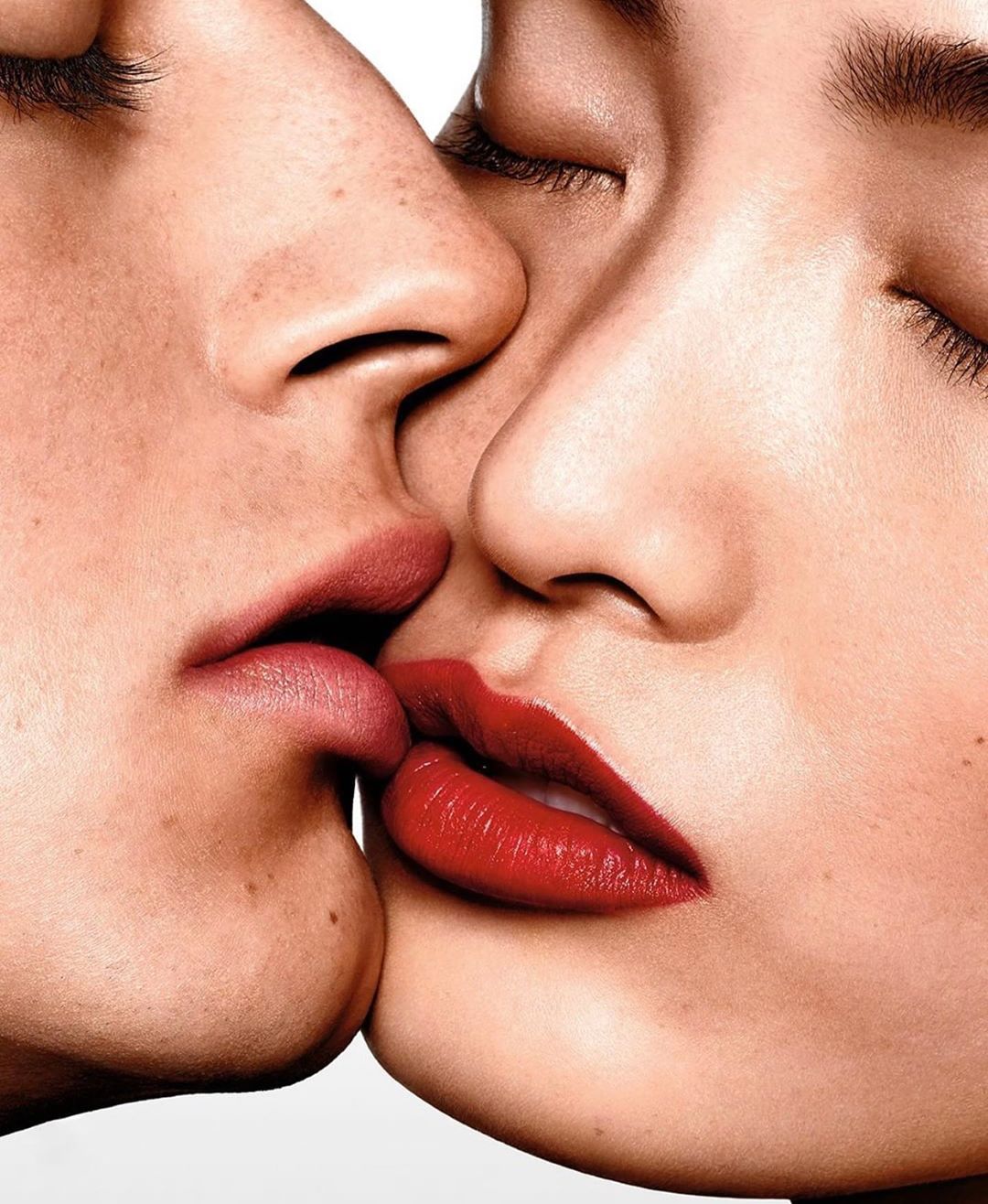Close-up studio portrait of two adults with fair skin kissing with red lips in a white background, makeup, beauty, and fashion.