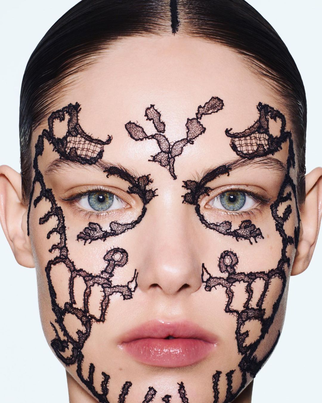 Close-up of an adult woman's face adorned with black lace patterns, set against a bright background in a beauty or fashion context.