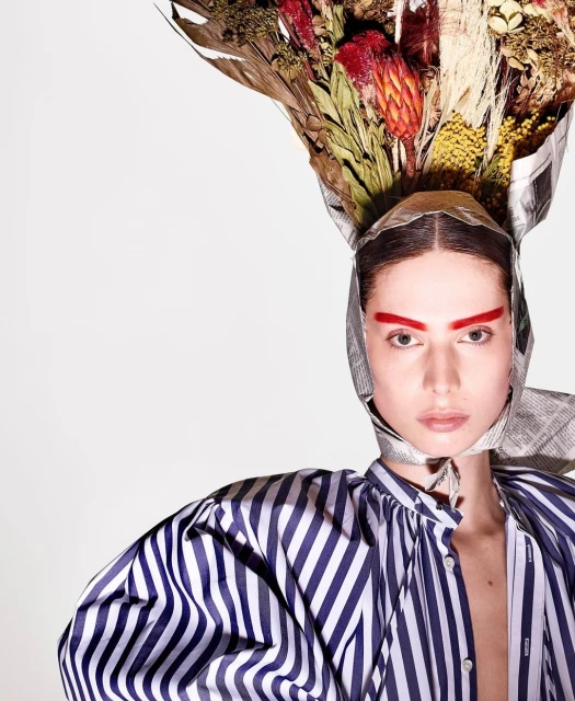 Adult woman with red eyebrows wearing striped shirt and headdress made from newspaper and dried flowers in a studio headshot.