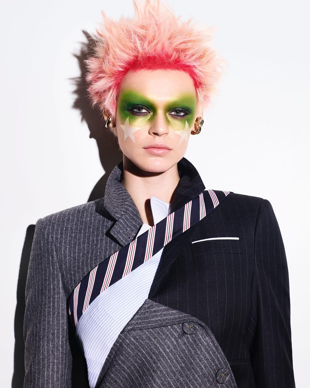 Eccentric fashion portrait: person with pink spiky hair, green makeup, star face paint, and a mismatched pinstripe blazer against a white background.