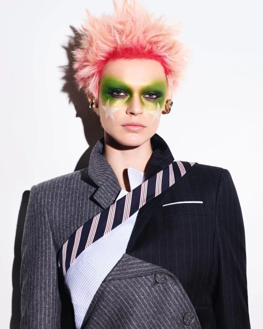 Eccentric fashion portrait: person with pink spiky hair, green makeup, star face paint, and a mismatched pinstripe blazer against a white background.
