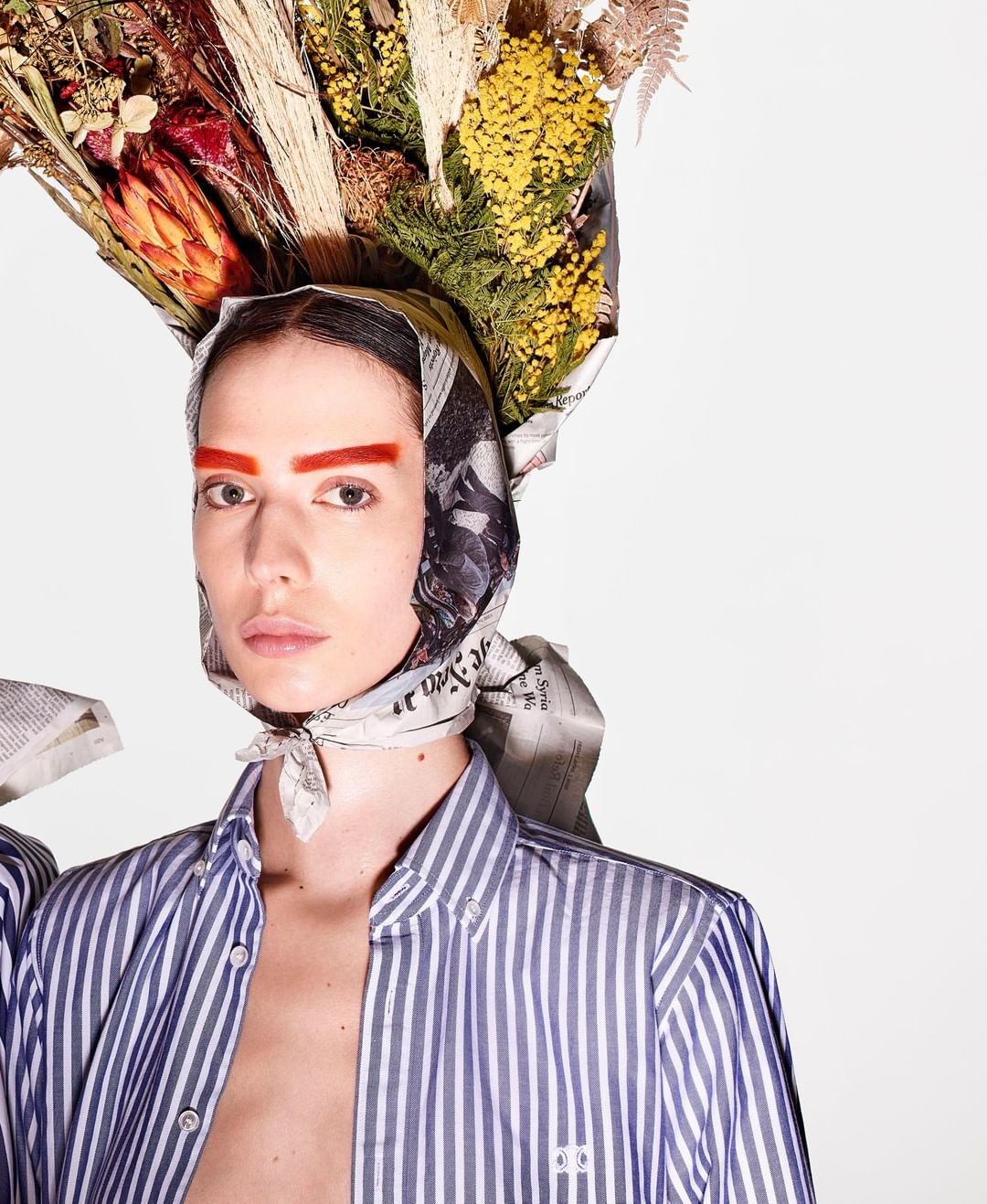 Striking editorial fashion portrait of a model with a newspaper headscarf and flowers on her head, against a clean, bright background.