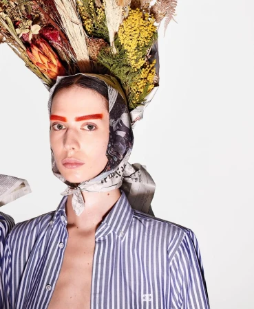 Striking editorial fashion portrait of a model with a newspaper headscarf and flowers on her head, against a clean, bright background.
